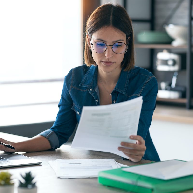 Shot of pretty young business woman working with computer while consulting some invoices and documents in the kitchen at home.